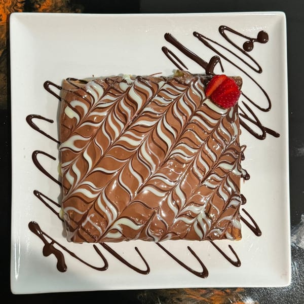 A square dessert with chocolate and white drizzle, topped with a strawberry, on a white plate.